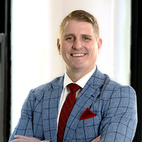 Young man in  checkered light blue suit with a burgundy tie standing in front of a window.