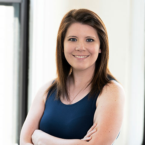 Young woman with medium length straight brown hair and blue shirt standing in front of the window